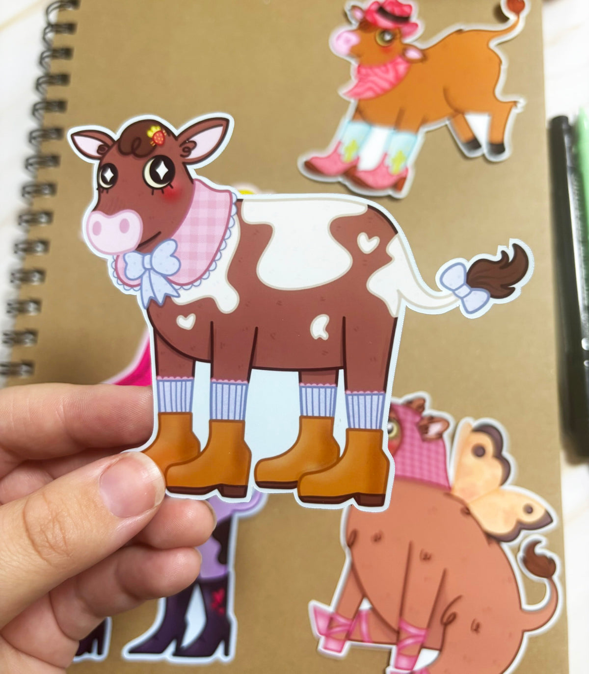 Brown-and-white cow sticker with a pink gingham collar, pale blue bows, striped socks and mustard-coloured boots.