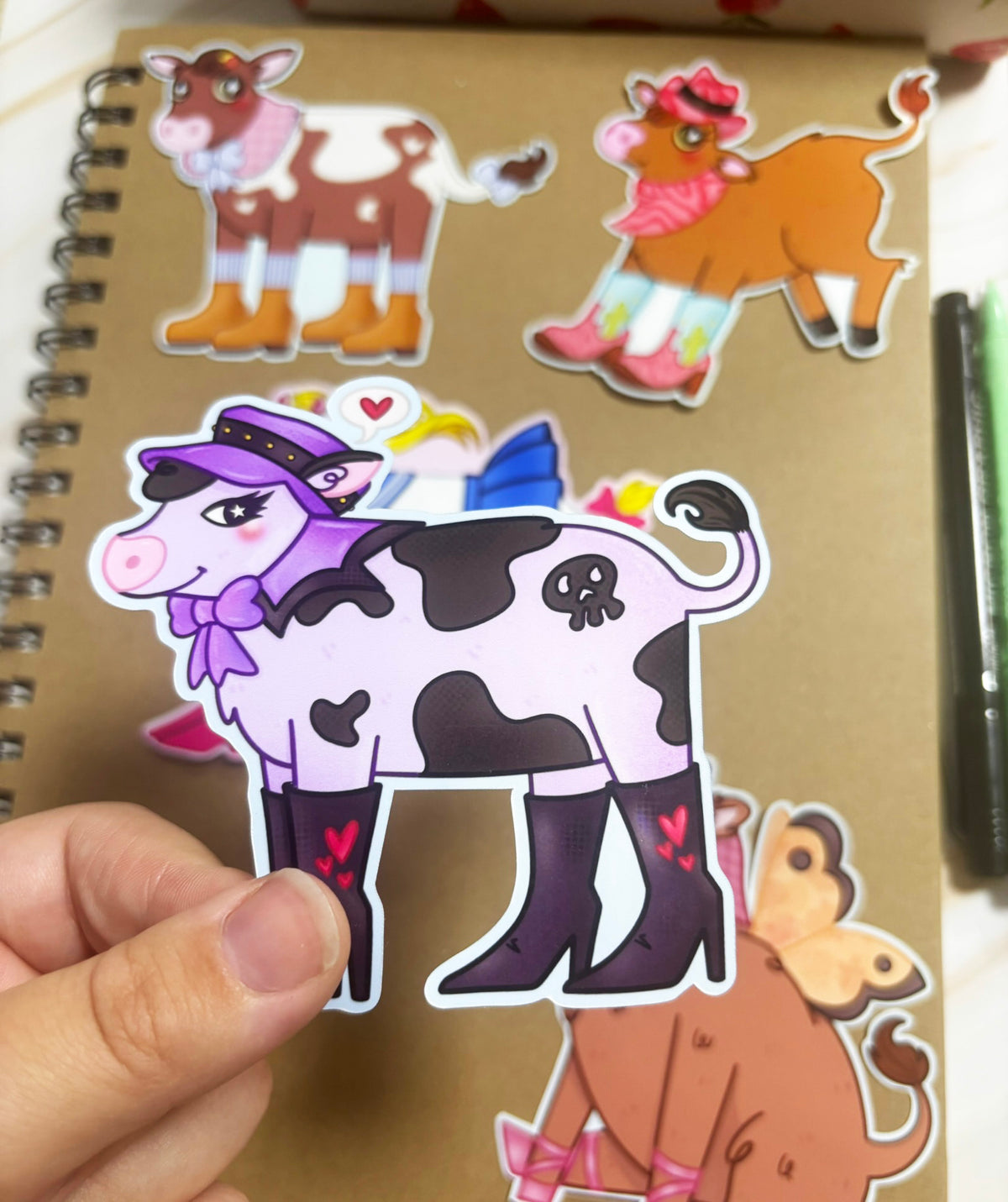 Pale lilac cow sticker with black patches, a skull-shaped marking, purple hat and bow, and black heeled boots with pink hearts.