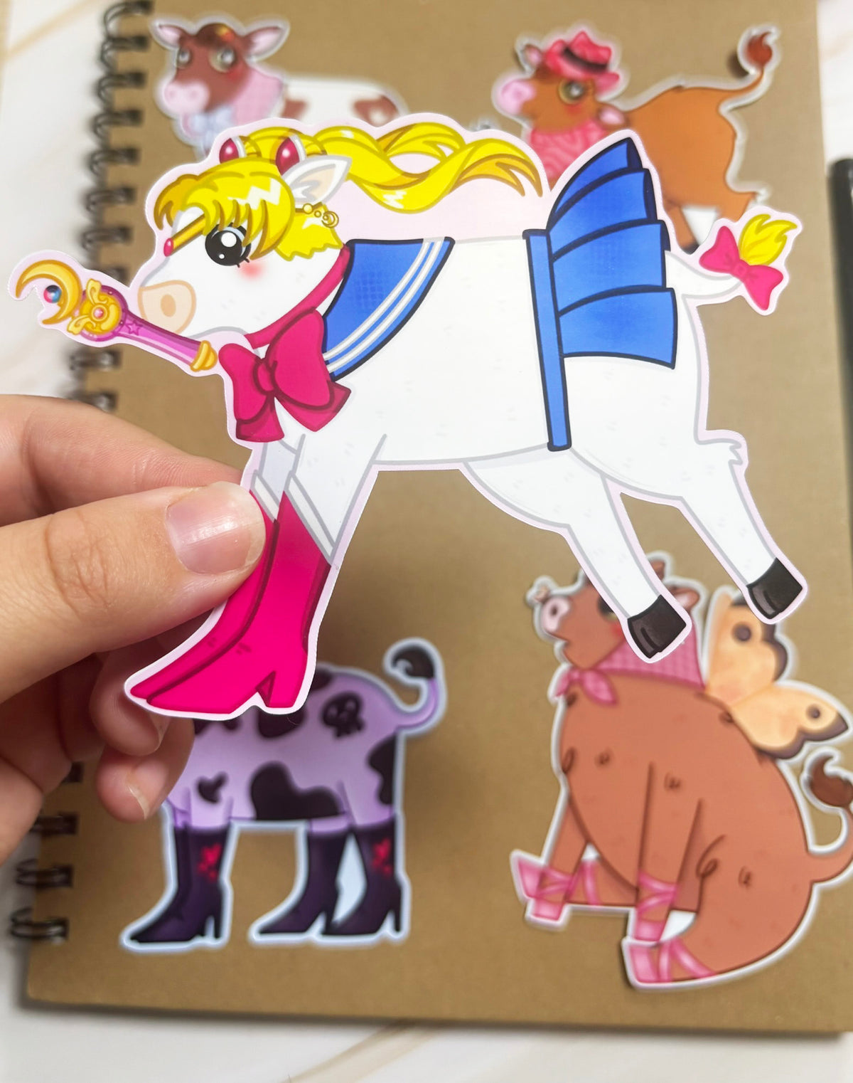 White cow sticker with yellow hair, a blue sailor-style outfit, pink bow, tall pink boots and a crescent-moon wand.