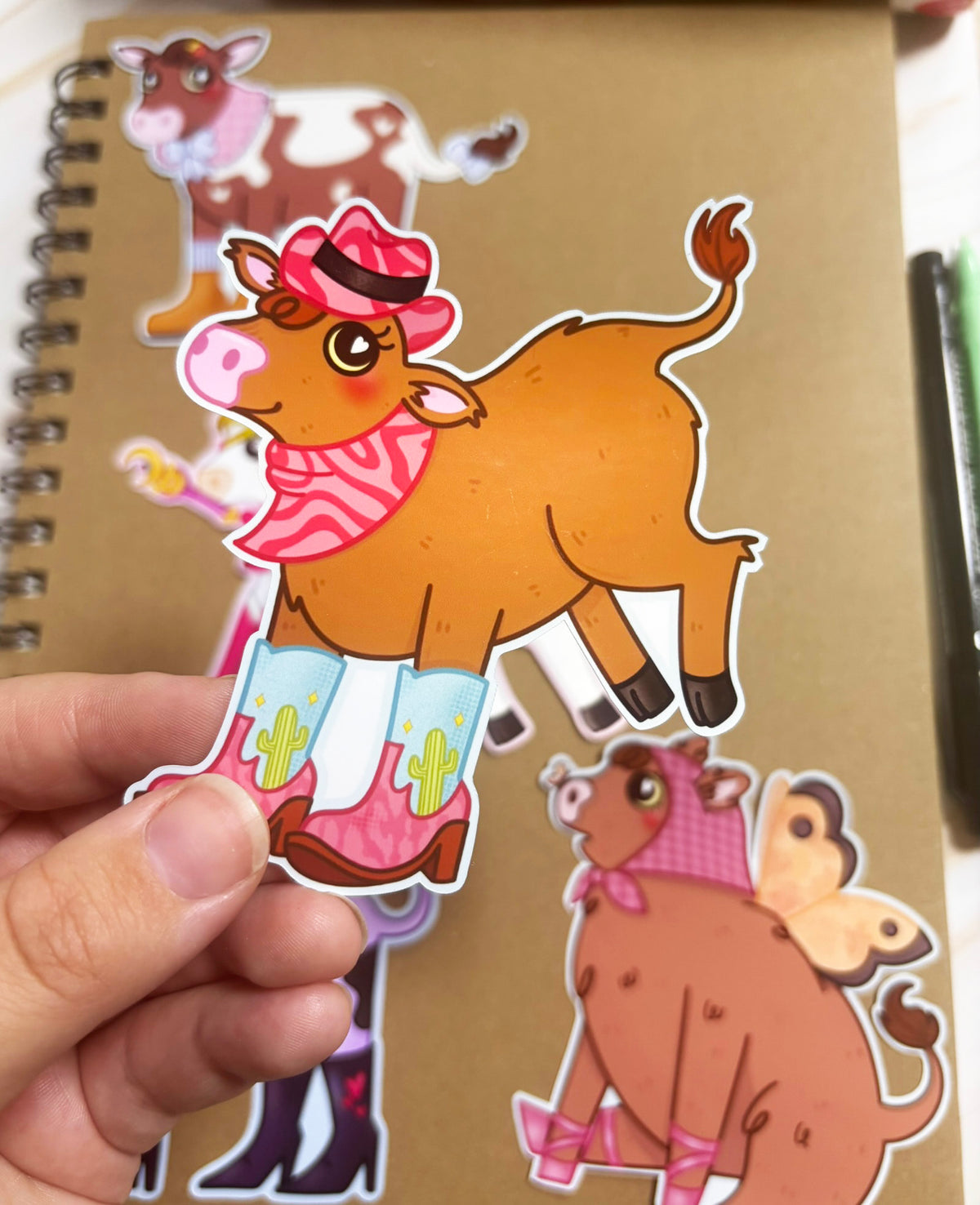 Brown cow sticker wearing a pink cowboy hat, pink bandana and pink-and-blue cowboy boots decorated with green cacti.